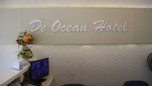 Un comptoir d’accueil à l’hôtel De Ocean, avec un écran, un arrangement floral et un panneau indicateur.
