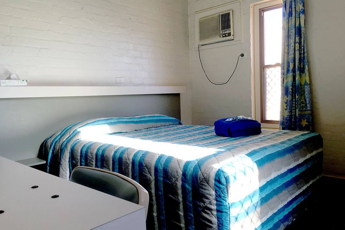 Ein Einzelbett mit gestreifter blau-weißer Bettdecke, ein Nachttisch, ein Stuhl, ein Fenster mit Vorhängen sowie eine wandmontierte Klimaanlage.