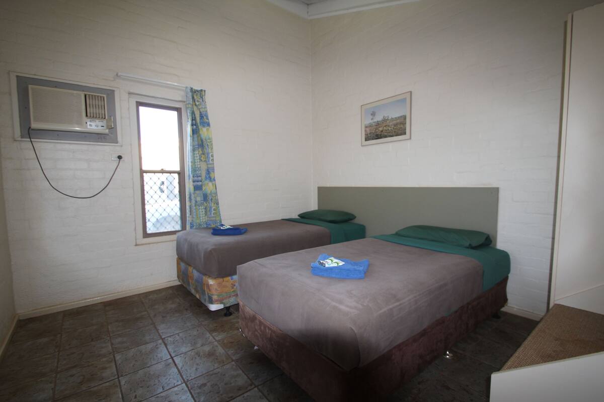 Ein Zimmer mit zwei Betten, einem Vorhangfenster, einer Klimaanlage und einem kleinen Bild an der Wand.