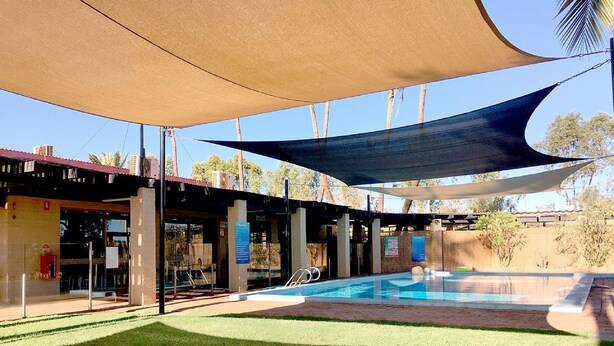Una piscina al aire libre con zona sombreada, rodeada de césped cuidado y un edificio con techo rojo.