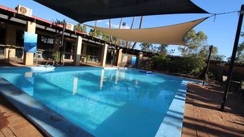 Une piscine dotée d’un espace ombragé, entourée d’une terrasse en briques et d’un bâtiment avec de grandes fenêtres et des unités de climatisation.