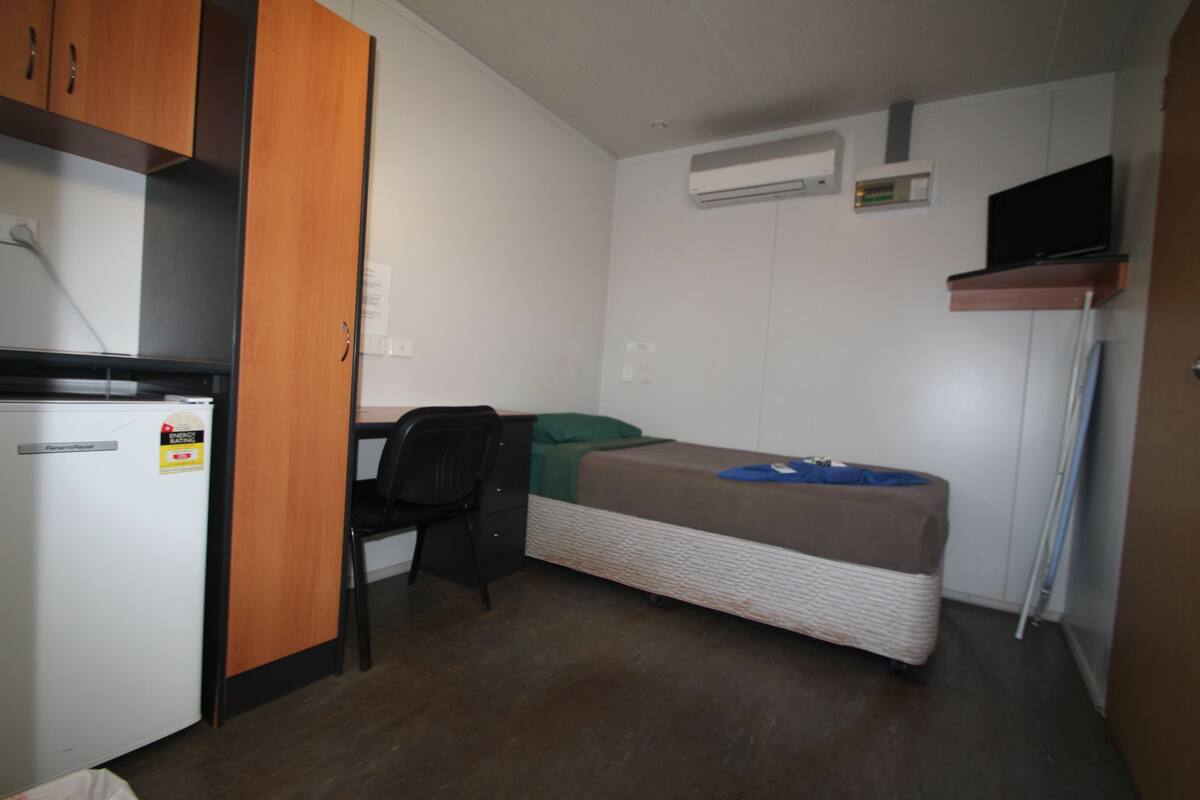 Ein kleiner Raum mit Bett, Schreibtisch, Stuhl, einer kleinen Küchenzeile und einem an der Wand befestigten Fernseher.