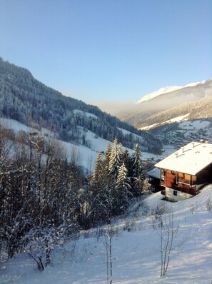 Eine moderne Chalet mit geneigtem Dach, gelegen an einem mit Schnee bedeckten Hang mit Blick auf ein Tal und entfernte Berge.