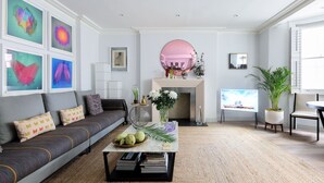 A living room with a grey sofa, a coffee table with books, a TV, and a fireplace with a pink mirror above it. - GREAT LOCATION -  Luxurious Knightsbridge Apartment - Steps from Harrods (London)