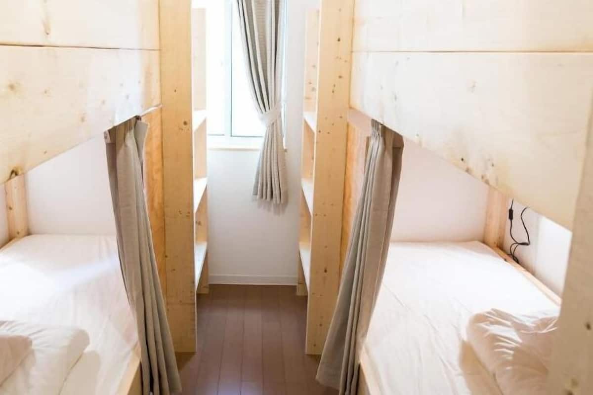 Ein schmaler Raum mit zwei Betten, Holzwänden, einem Fenster mit Vorhängen und einer sichtbaren Steckdose.