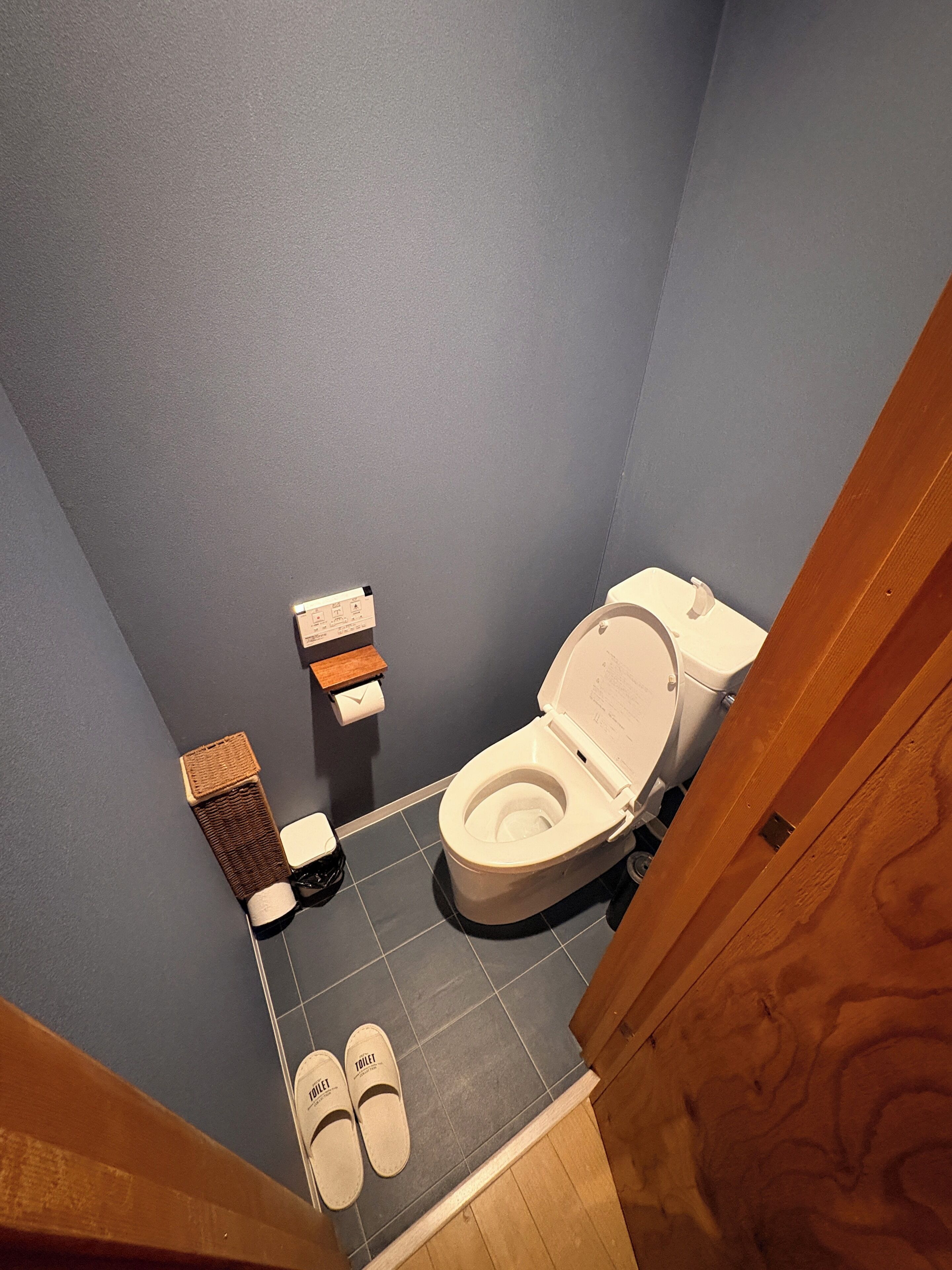 A small bathroom with a toilet, a trash bin, and a pair of slippers on the floor.