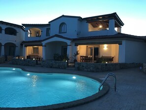 Una gran piscina con borde de piedra, un patio con muebles de exterior y un edificio de varios pisos con ventanas arqueadas y techo de tejas.