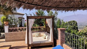 Zona de estar al aire libre con techo de paja, cama con dosel de madera y sillón azul.