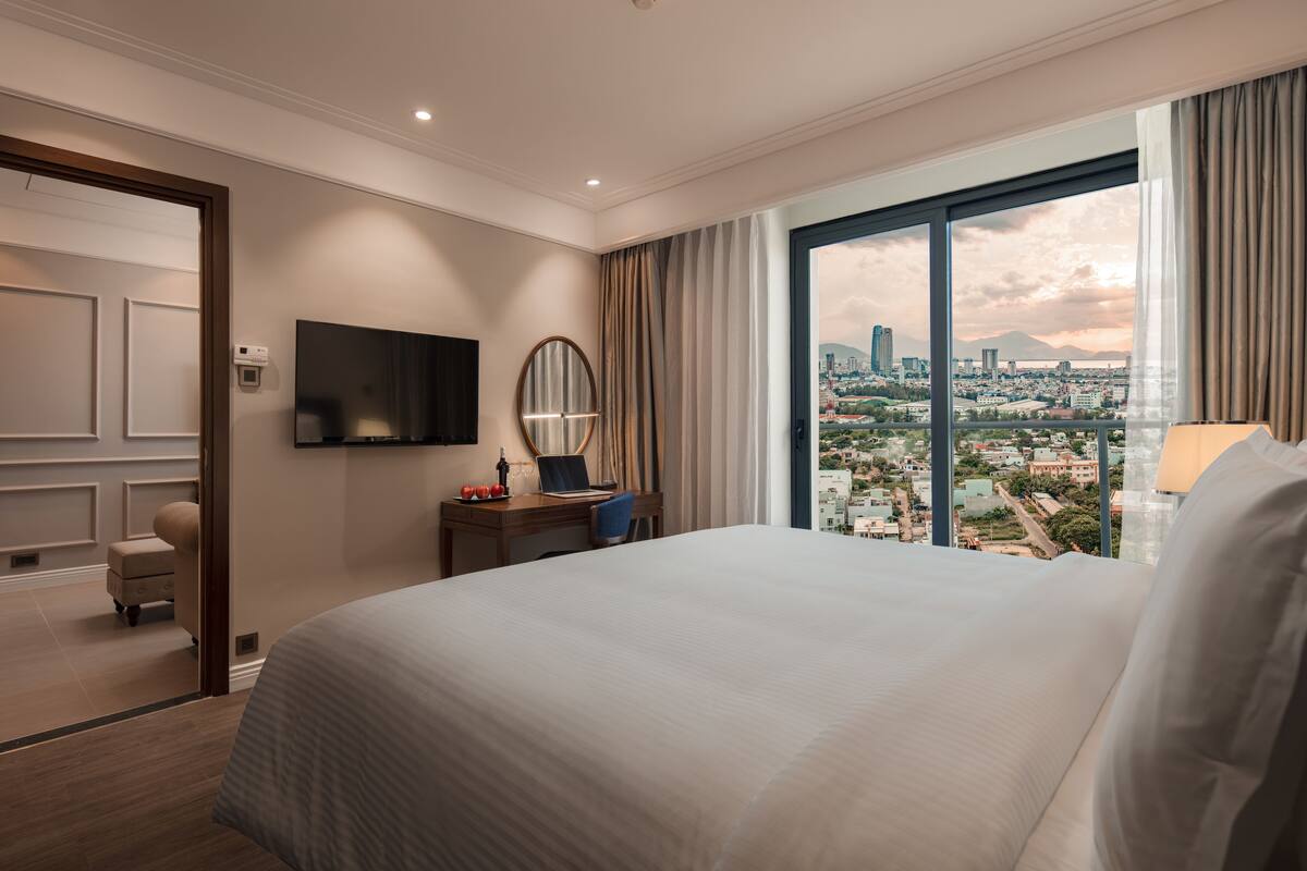 Une chambre d’hôtel avec un grand lit, un bureau avec un ordinateur portable, une télévision et une vue sur le paysage urbain par une grande fenêtre.