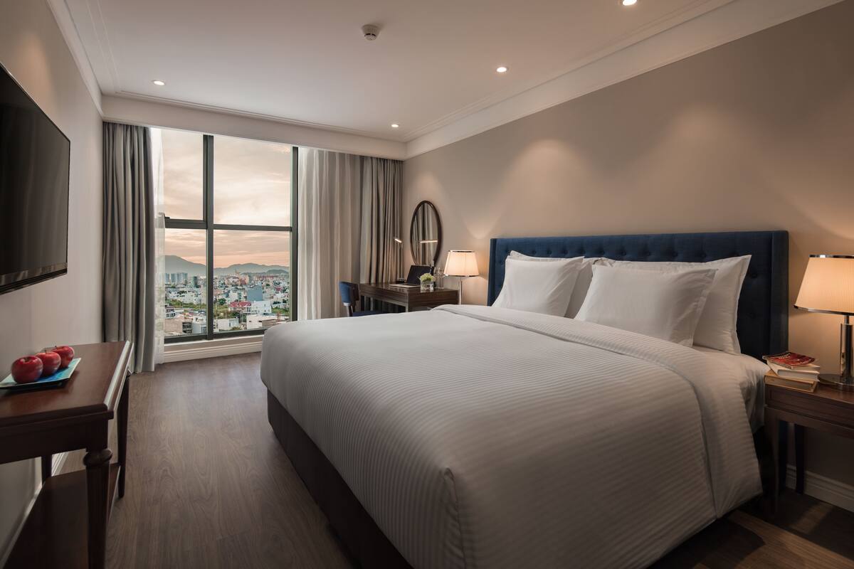 Une chambre d’hôtel avec un grand lit, un bureau avec une télévision et une vue sur le paysage urbain par une grande fenêtre.