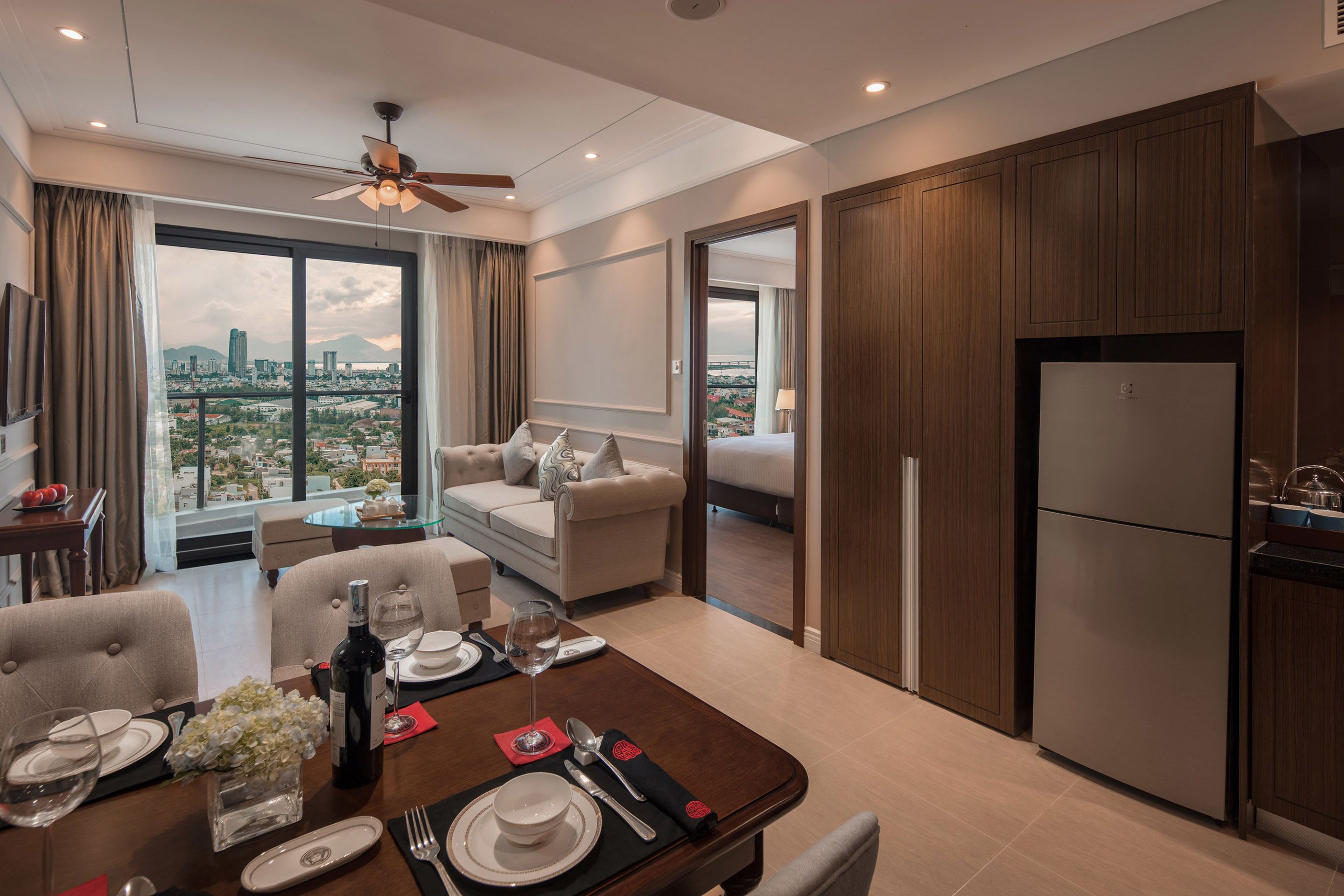 A modern hotel room with a dining area, a sofa, a ceiling fan, and a view of the cityscape.