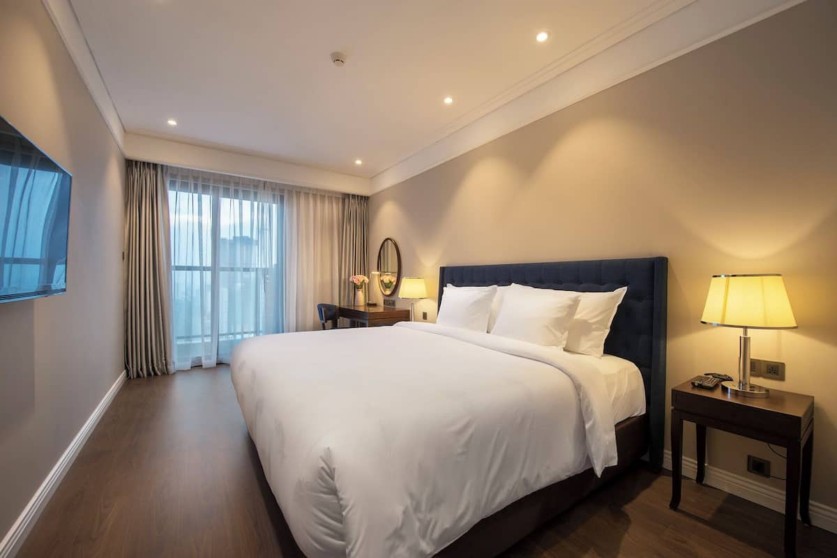 Une chambre d’hôtel avec un grand lit, un bureau, une chaise, une lampe et une fenêtre avec des rideaux.