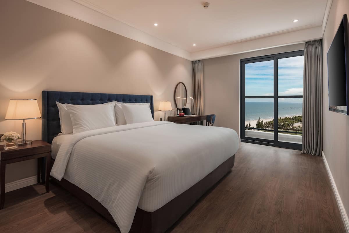 Une chambre d’hôtel avec un grand lit, une table de chevet avec une lampe, un bureau avec un miroir et une vue sur l’océan par une grande fenêtre.