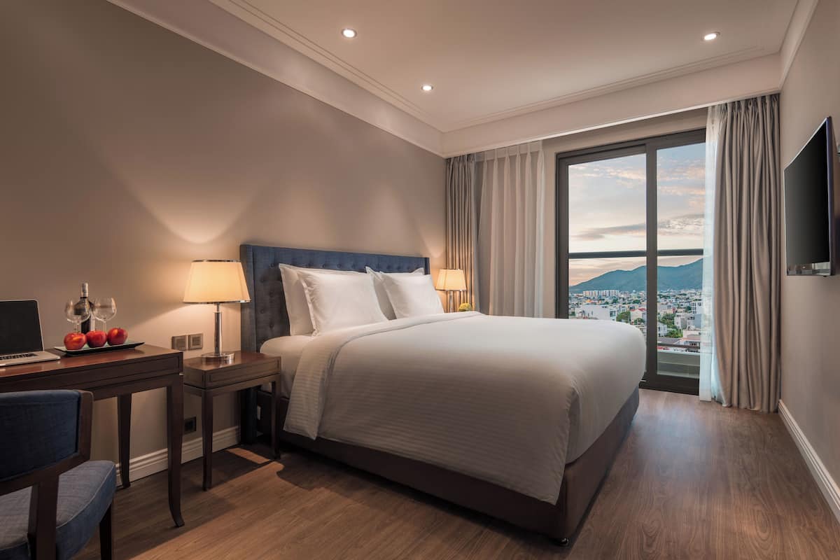 Une chambre d’hôtel avec un grand lit, un bureau avec un ordinateur portable et des verres à vin, et une vue sur le paysage urbain à travers une grande fenêtre.