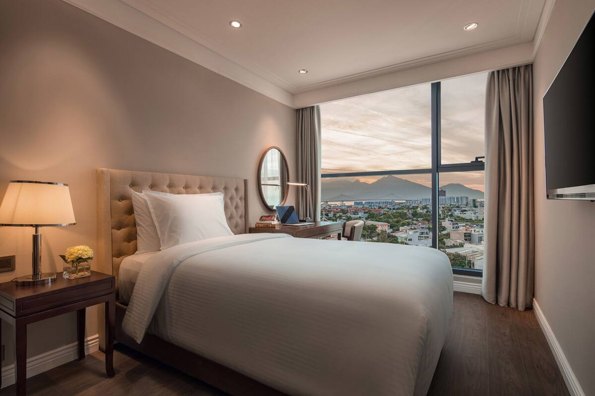 Une chambre d’hôtel avec un grand lit, une table de chevet, une lampe, un miroir et une vue sur le paysage urbain, visible à travers de grandes fenêtres.