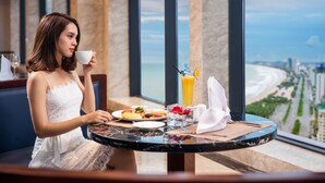 Une femme vêtue d’une robe blanche est assise à une table où se trouvent un repas et une boisson. Elle a devant elle une vue sur la ville et la plage.