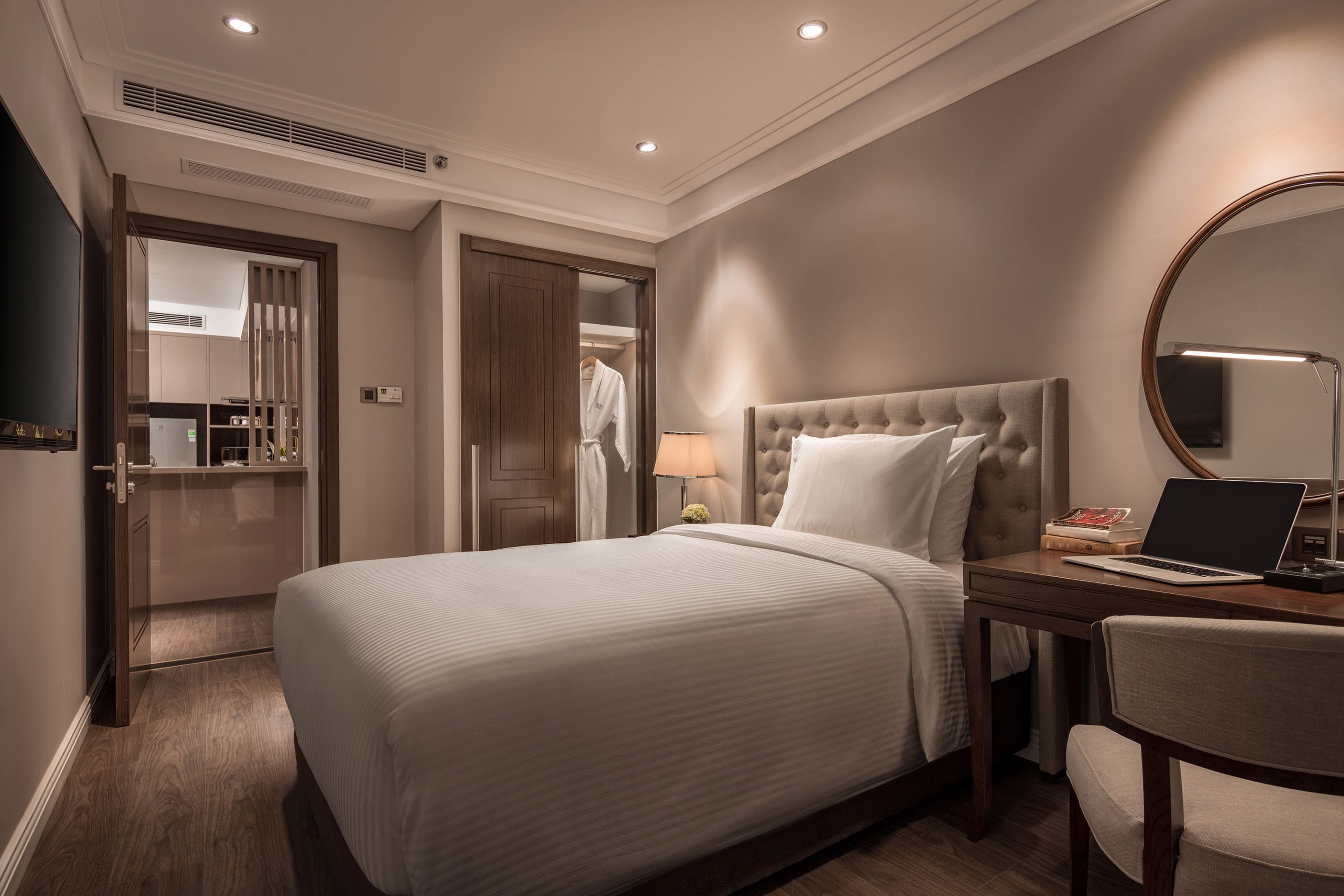 A hotel room with a large bed, a desk with a laptop, a round mirror, and a wardrobe.