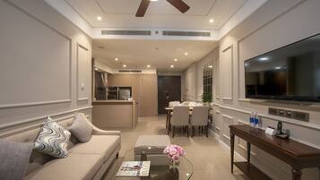 A modern living room with a sofa, coffee table, and a TV. There is a dining area with chairs and a table, and a kitchen in the background.