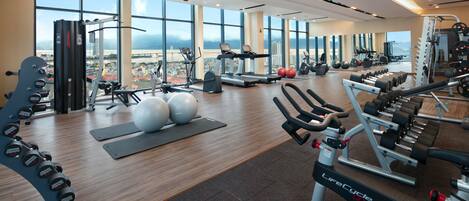 Un gimnasio con máquinas de cardio, pesas y pelotas de ejercicio, con amplias ventanas y vistas a un paisaje urbano.