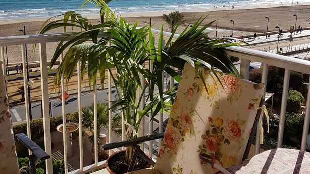 A balcony with a view of the beach, featuring red umbrellas, potted plants, and floral cushions. - BEAUTIFUL APARTMENT VACATION RENTAL PLAYA SAN JUAN ALICANTE (san juan alicante)