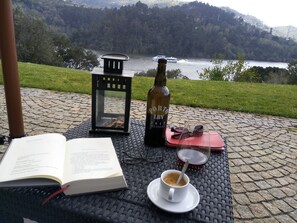 An open book, a bottle of Porto wine, a pair of glasses, and a cup of coffee on a table with a view of a lake and hills. - Douro Valley With Family (Paços de Gaiolo)