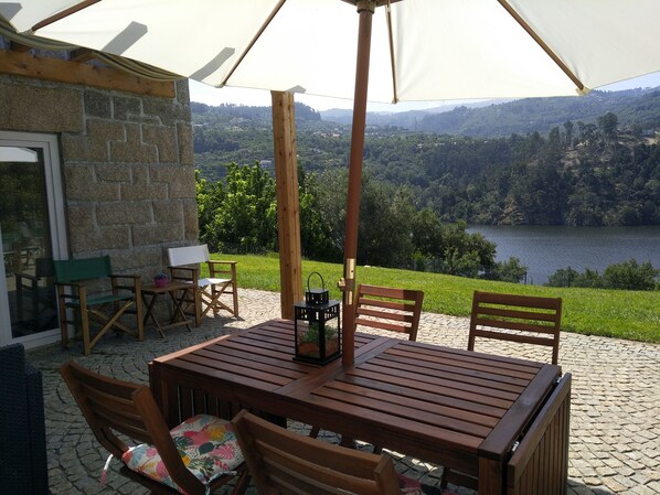 An outdoor patio with a wooden table and chairs, a white umbrella, and a view of a lake and hills.
