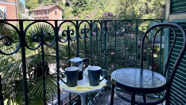Un balcón con barandilla metálica, una mesa pequeña con una silla, y una bandeja con dos tazas y una tetera.