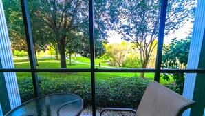 A view through a window with a table and chair, overlooking a green park with trees and a pathway.