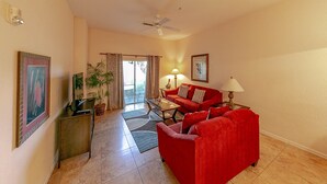 A living room with a red sofa, a coffee table, and a lamp. There is a ceiling fan, a plant, and a door leading to another room.