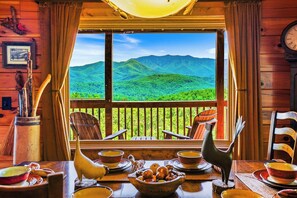 A wooden cabin with a dining table set for two, a view of green mountains through a window, and rustic decor including a wooden chair and a wall clock.