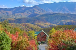 A cabin with a green roof and a red exterior, surrounded by autumn foliage and mountains in the background.