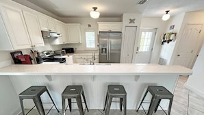 A kitchen with white cabinets, a stainless steel refrigerator, a microwave, and a sink. There are four stools in front of the counter. - Beautiful, well-appointed three-bedroom beach house 300 yards from the beach. (St Augustine)