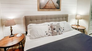 Quarto com cama, duas mesas de cabeceira, um criado-mudo, um quadro na parede e piso de madeira.