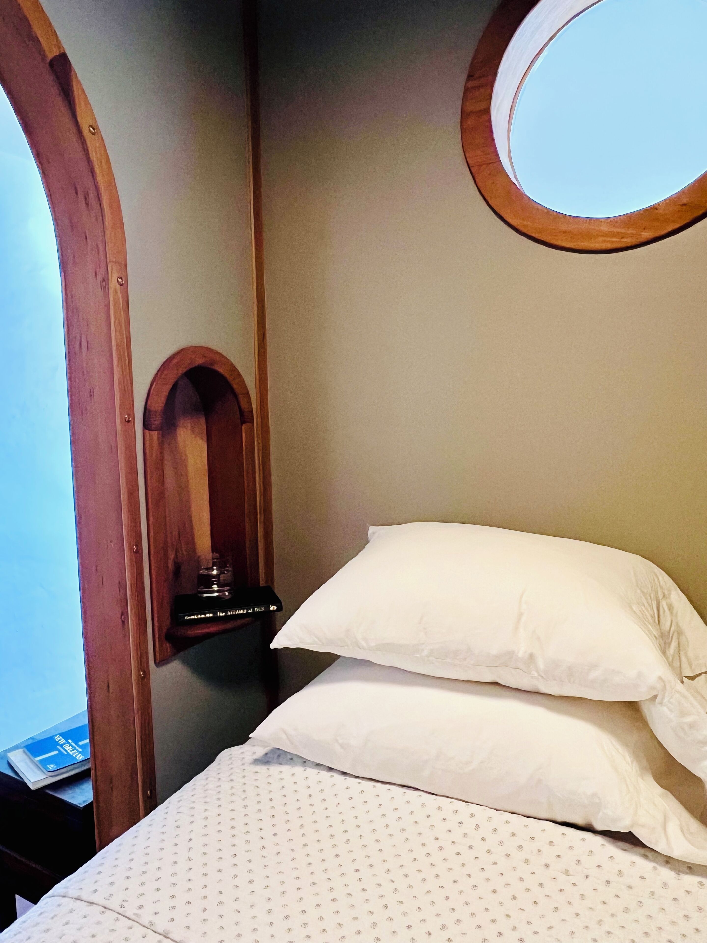 A bedroom with a bed, two pillows, a small wooden shelf with a book, and a round mirror on the wall.