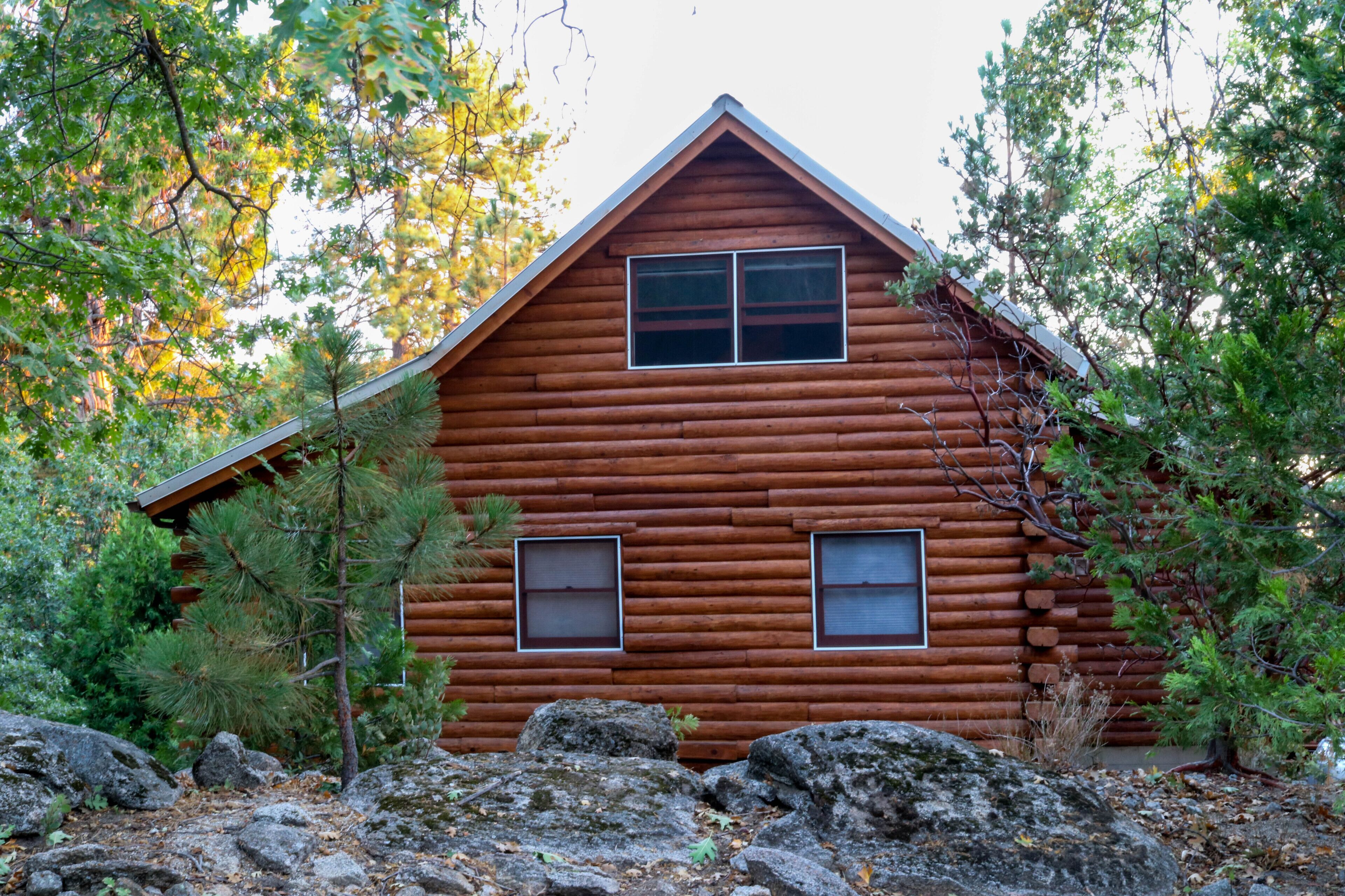 A log cabin with two windows and a triangular roof, surrounded by trees and rocks.