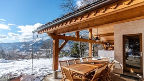 Une terrasse en bois avec une table et des chaises, d’où l’on peut admirer un paysage enneigé avec des montagnes en arrière-plan.