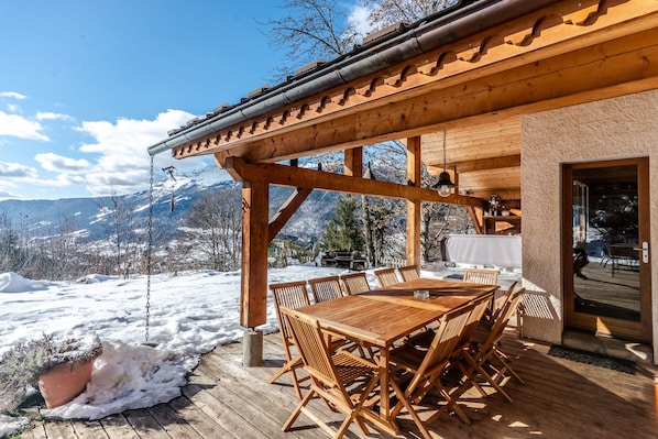 A wooden deck with a table and chairs, overlooking a snowy landscape with mountains in the background.