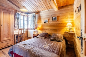 A wooden cabin bedroom with a bed, two bedside lamps, a chair, a window with curtains, and a wooden wardrobe.