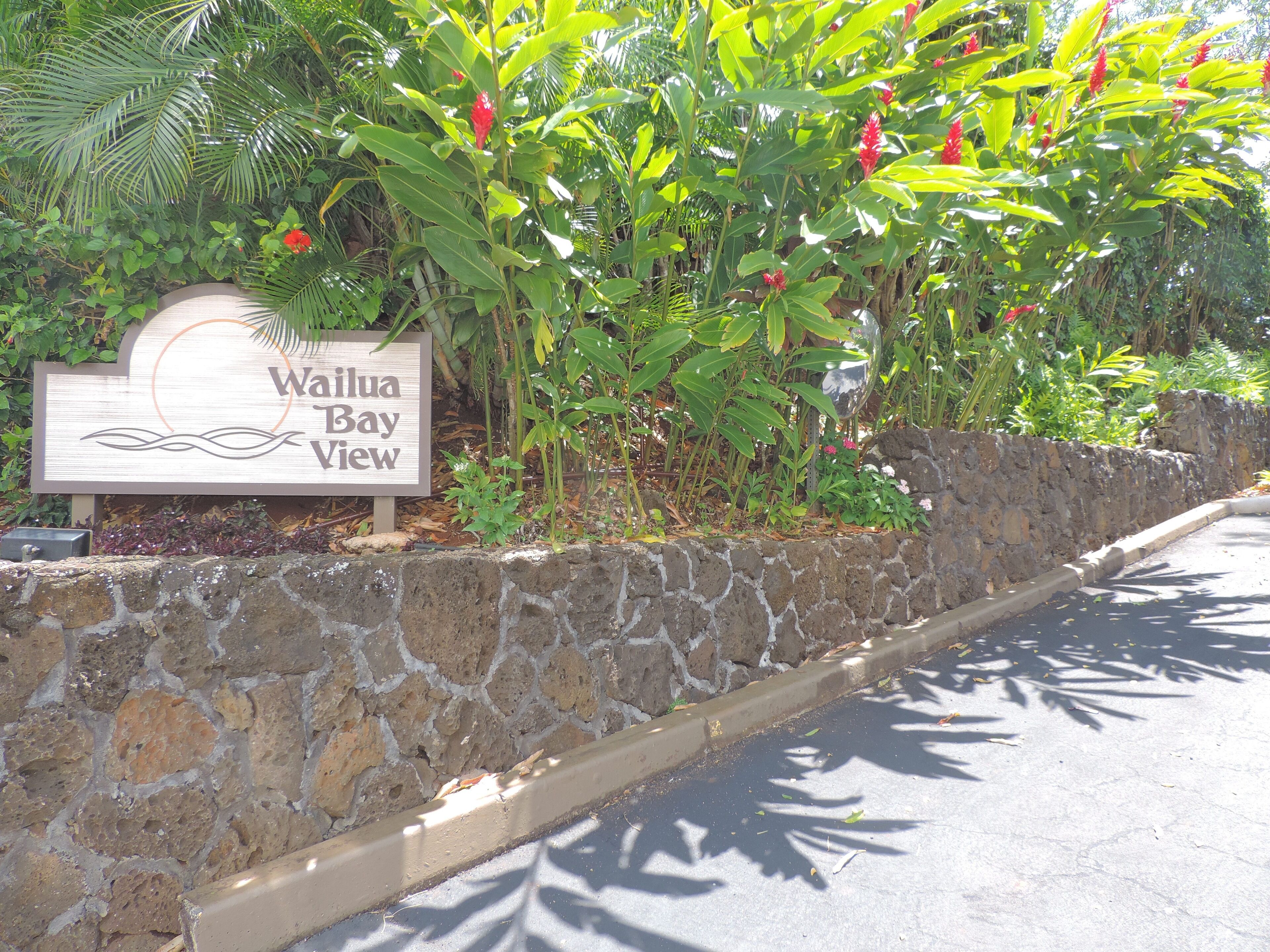 A sign for Waikoloa Bay View, surrounded by lush greenery and a stone wall.
