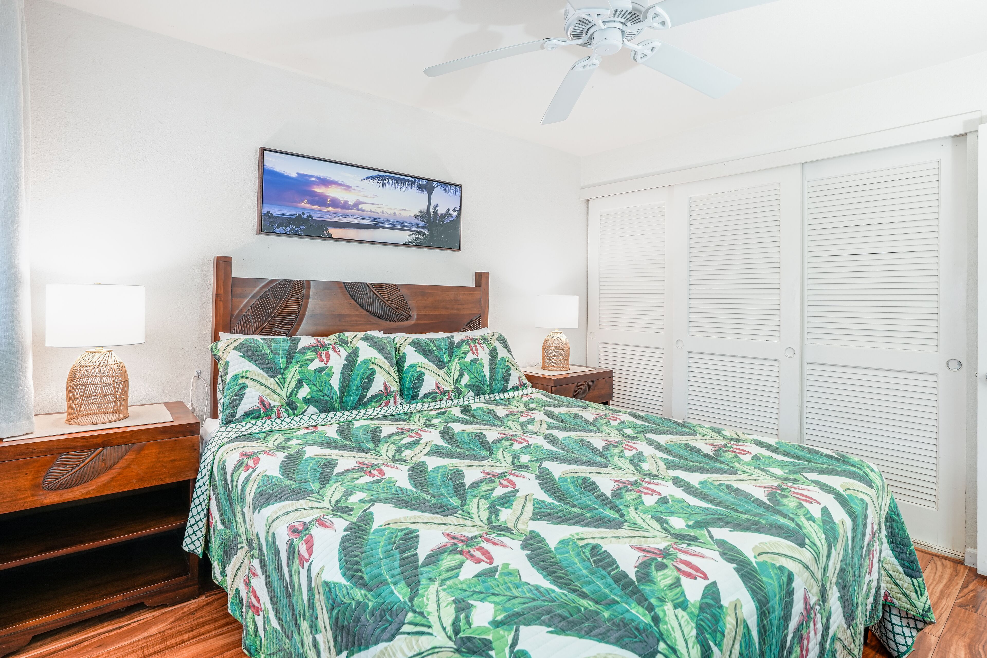 A bedroom with a bed, wooden nightstands, a ceiling fan, a wall-mounted TV, and a door with louvered shutters.