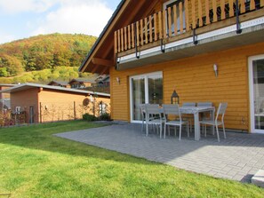En træbygning med balkon, en terrasse med bord og stole, og udsigt til en bakketop.