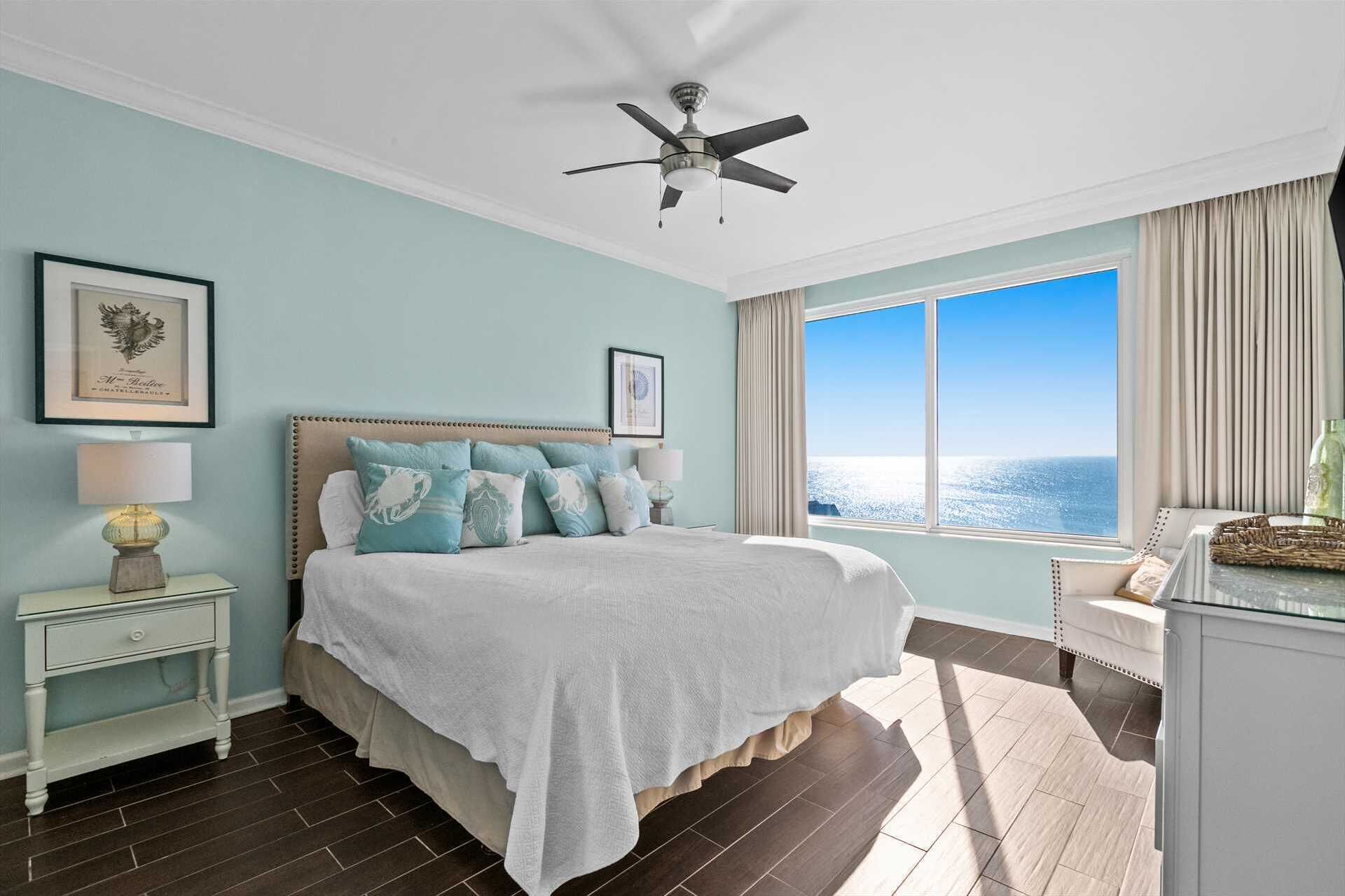 A bedroom with a bed, a nightstand, a lamp, a ceiling fan, a window with a view of the ocean, and a framed picture on the wall.