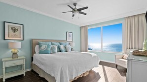 Un dormitorio con cama, mesita de noche, lámpara, ventilador de techo, ventana con vista al océano y un cuadro en la pared.