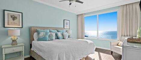 A bedroom with a bed, a nightstand, a lamp, a ceiling fan, a window with a view of the ocean, and a framed picture on the wall.