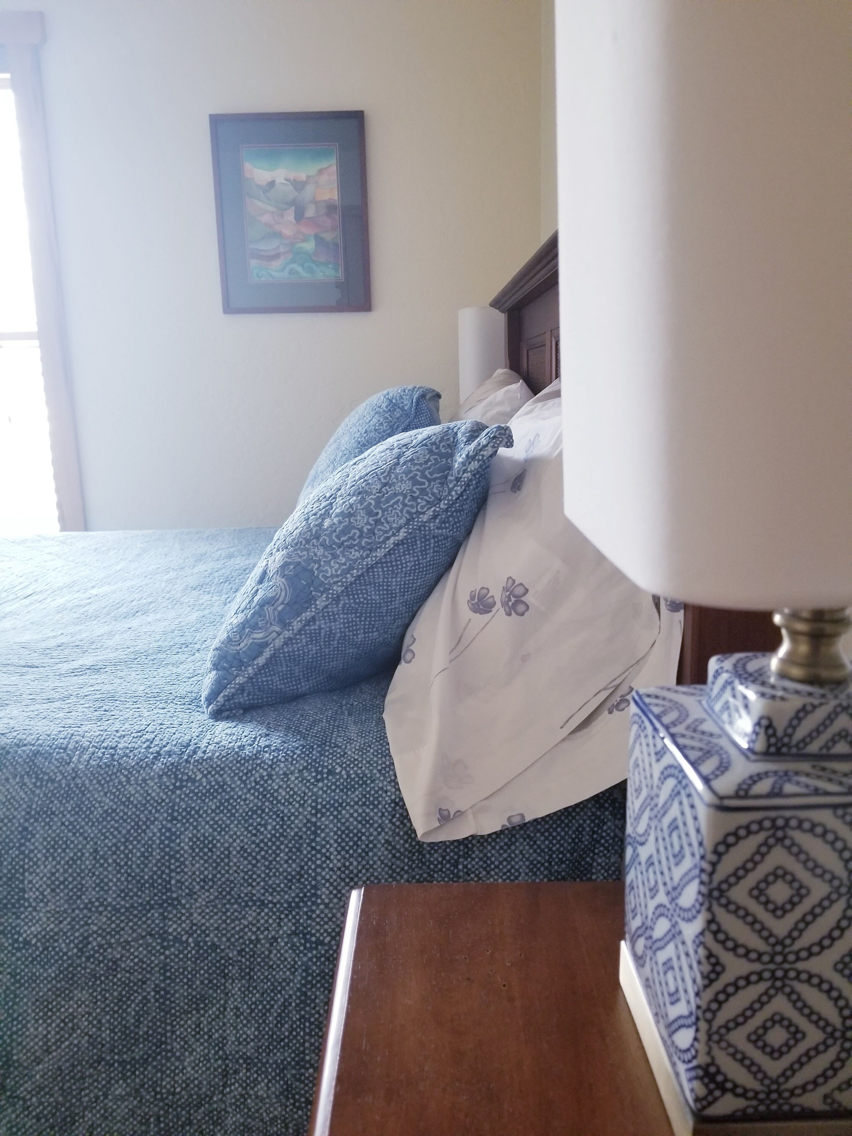 Ein ordentlich gemachtes Bett mit blau-weißer Bettwäsche, ein gerahmtes Bild an der Wand und ein Nachttisch mit einer Lampe mit Muster.
