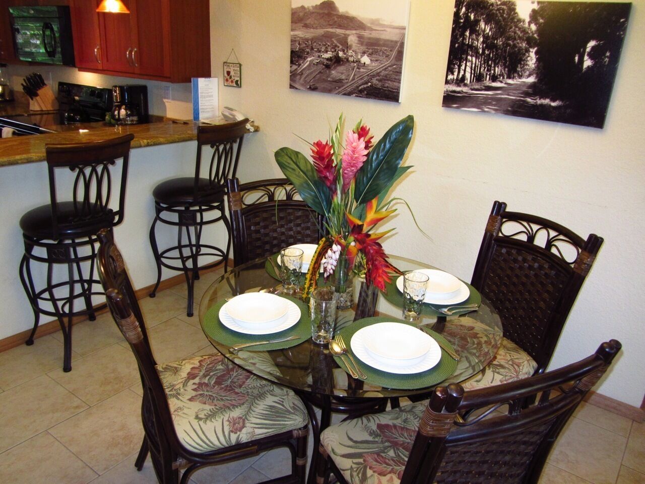 A dining area with a round glass table set for four, surrounded by dark wooden chairs with floral upholstery. A centerpiece with flowers and a small vase is on the table.