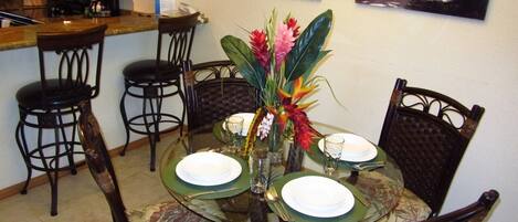 A dining area with a round glass table set for four, surrounded by dark wooden chairs with floral upholstery. A centerpiece with flowers and a small vase is on the table.