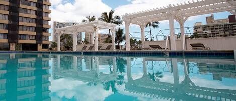 A swimming pool with a clear blue water surface, surrounded by lounge chairs and a white pergola structure.