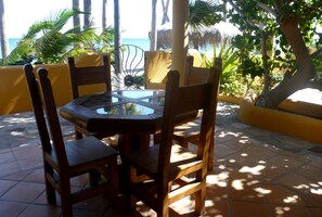 A round wooden table with chairs, surrounded by yellow walls and palm trees, overlooking a beach.