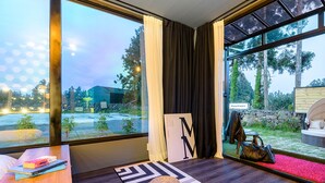 Quarto moderno com amplas janelas, uma mesa de madeira, um tapete e vista para área externa com parede de pedra e vegetação.